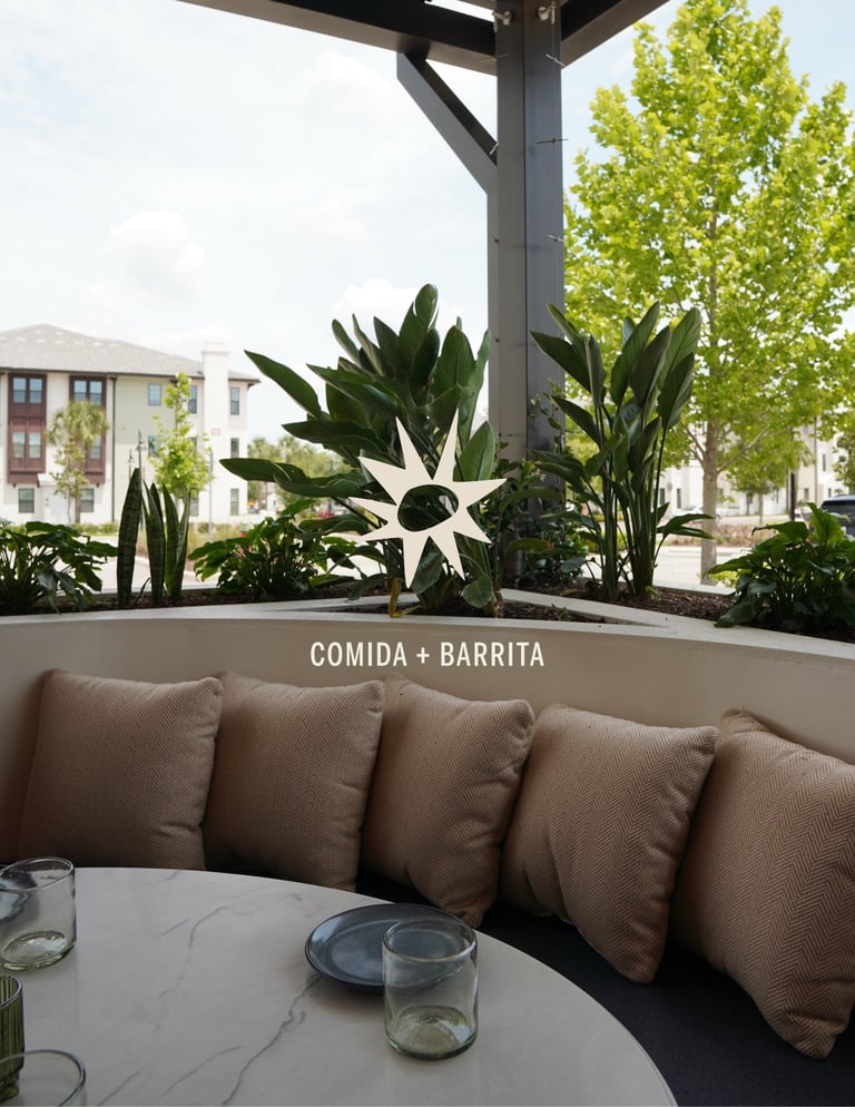 Rooftop restaurant patio with brown cushioned seating, marble table, potted plants, and city buildings visible in background