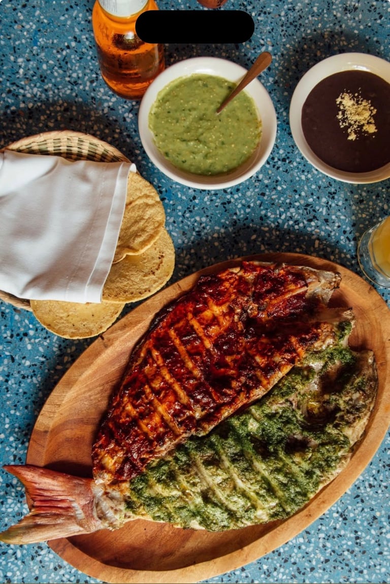 Whole roasted fish on wooden plate with green and red salsa, corn tortillas, and beverages on blue surface
