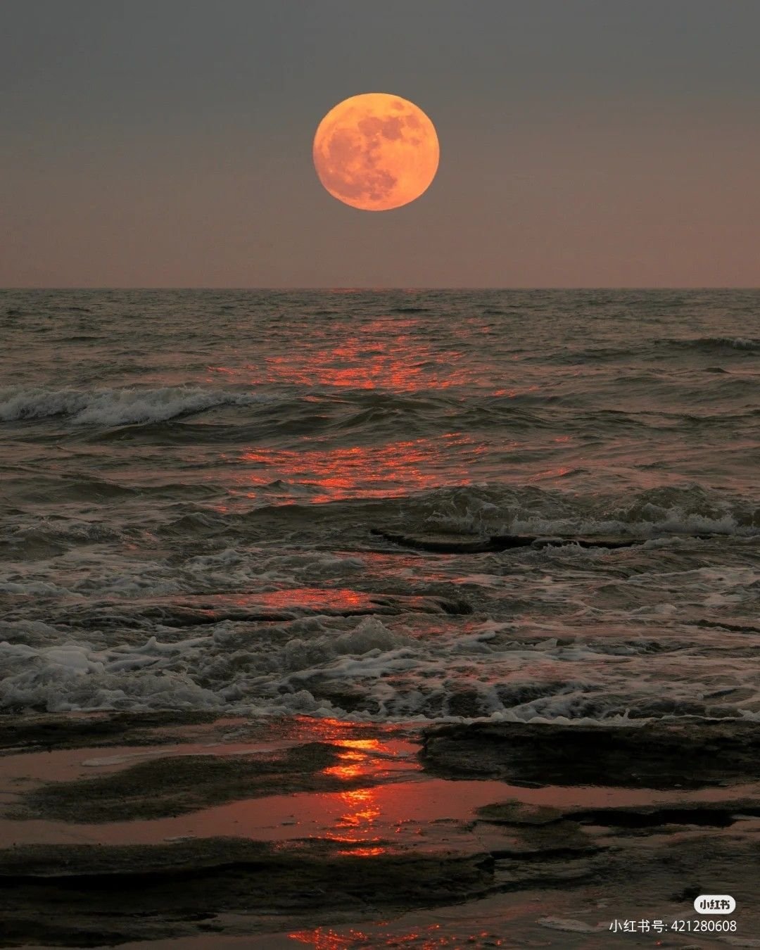 Large orange full moon rising over turbulent ocean waves at dusk, with moonlight reflecting on the dark water.